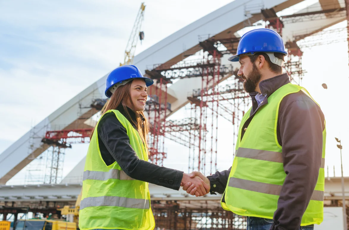 Two construction workers in safety vests and hard hats shake hands on-site, symbolising partnership and collaboration in infrastructure projects.